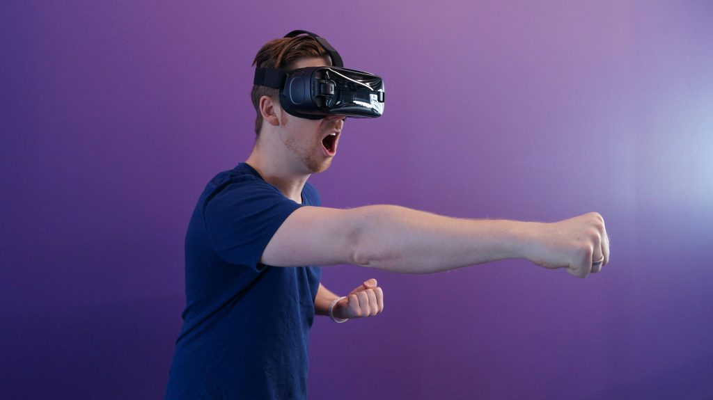 Caucasian man immersed in virtual reality using VR headset, making a punching gesture against a purple backdrop.